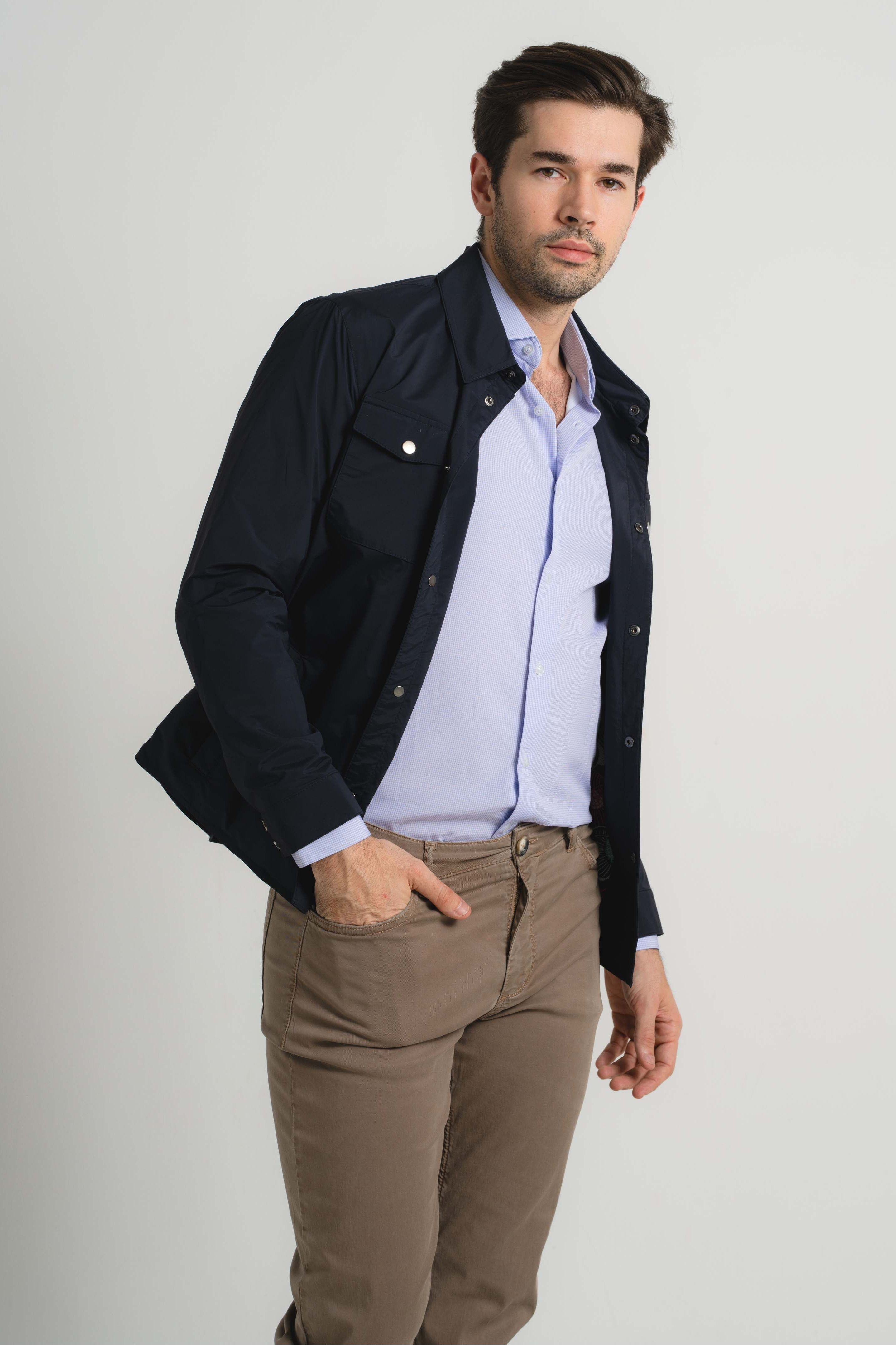 Man wearing a dark jacket, light shirt, and beige pants on a white background